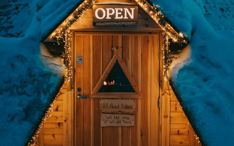 Eine gemütliche, schneebedeckte Hütte in Rovaniemi hat einen dreieckigen, warm beleuchteten Eingang. Auf den Schildern steht „Open“ und festliche Lichterketten umrahmen die Tür.