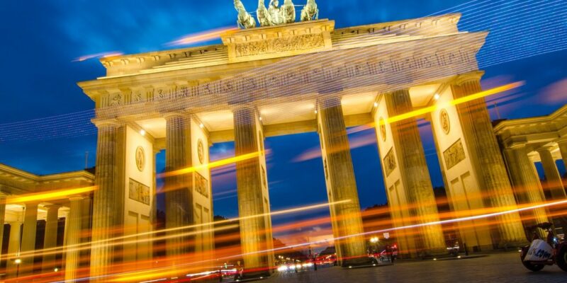 Das Bild zeigt das beleuchtete Brandenburger Tor in Berlin bei Nacht. Lichtspuren vorbeifahrender Fahrzeuge erzeugen im Vordergrund dynamische Streifen in Orange, Gelb und Blau und verleihen diesem ikonischen Berliner Wahrzeichen ein Gefühl von Bewegung.