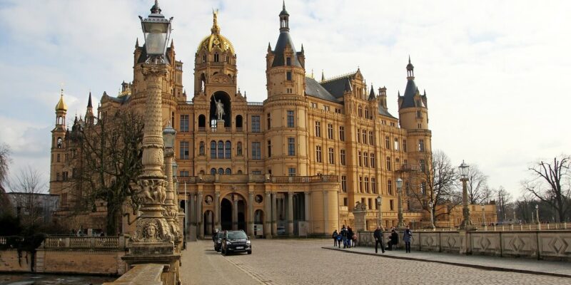 Ein großes, reich verziertes Schloss mit einer goldenen Kuppel und zahlreichen Türmchen vor einem wolkigen Himmel. Ein gepflasterter Weg führt zum Eingang. Menschen gehen spazieren und ein Auto parkt in der Nähe. Blattlose Bäume rahmen die Szene ein.