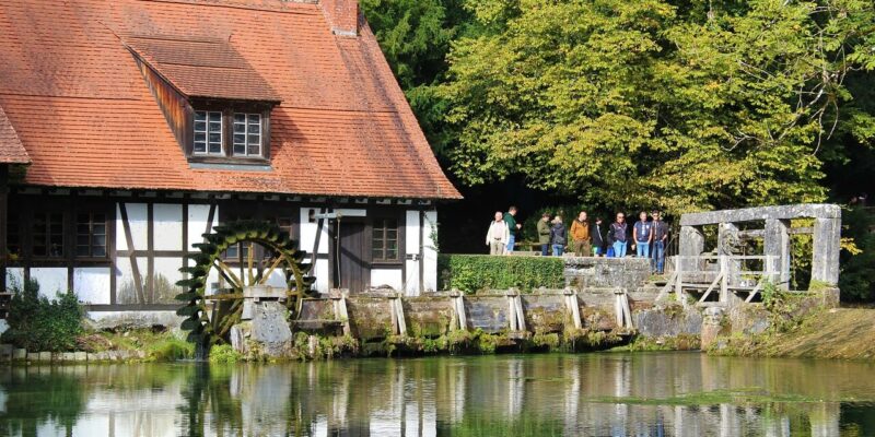 Eine malerische Szene zeigt ein Fachwerkhaus mit rotem Dach neben einer Wassermühle, die an den bezaubernden Blautopf in Baden-Württemberg erinnert. Auf einer nahe gelegenen Brücke steht eine Gruppe von Menschen, umgeben von üppigem Grün. Ihre Spiegelbilder sind im ruhigen Wasser darunter sichtbar.