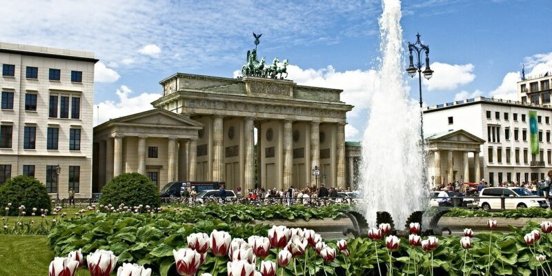 Ein lebendiger Garten mit roten und weißen Tulpen im Vordergrund, einem großen Brunnen und dem historischen Brandenburger Tor in Berlin, mit einem blauen Himmel und vereinzelten Wolken darüber.
