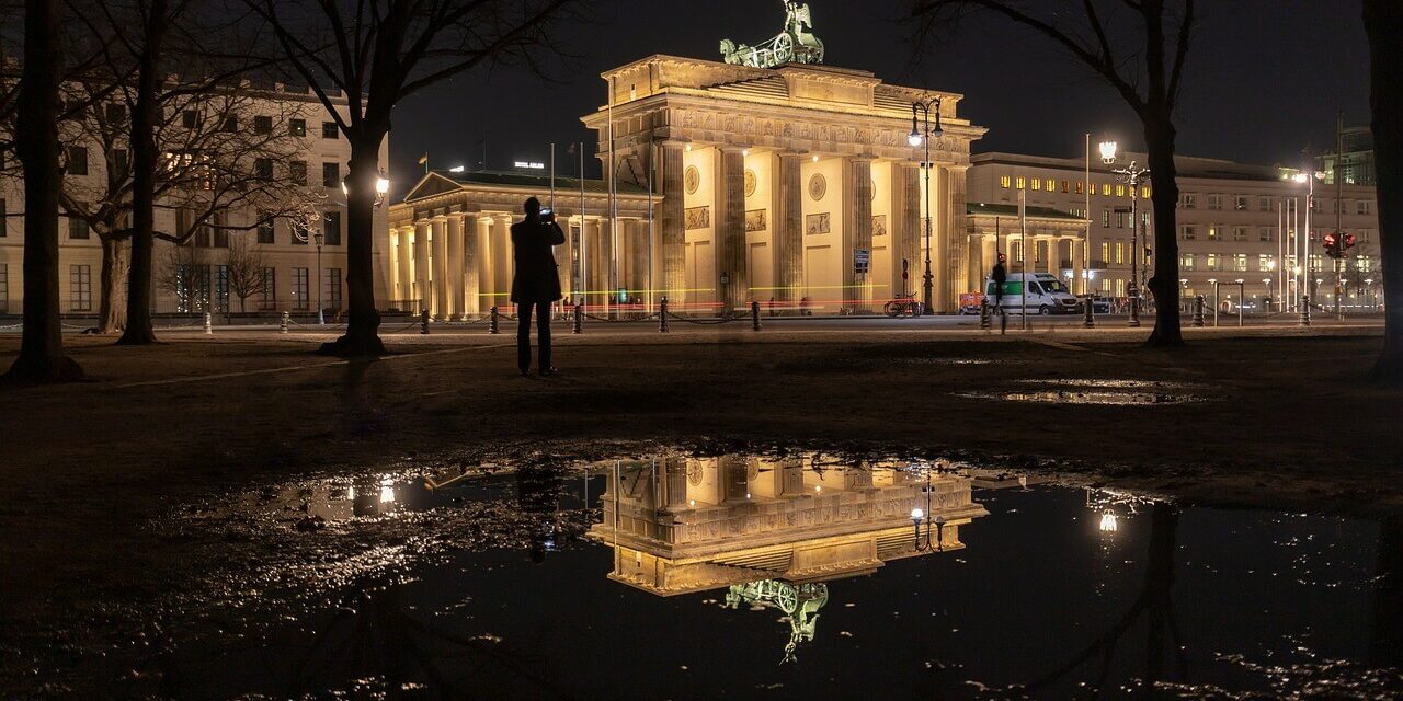 Das Nachtbild zeigt das Brandenburger Tor in Berlin, wunderschön beleuchtet und in einer großen Pfütze reflektiert. Silhouetten von Bäumen und einer einsamen Gestalt sind in der dunklen Umgebung subtil zu erkennen und verleihen der ruhigen Szene Tiefe.