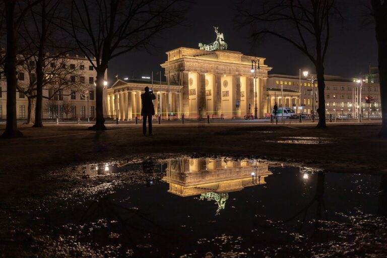 Brandenburger Tor Das Nachtbild zeigt das Brandenburger Tor in Berlin, wunderschön beleuchtet und in einer großen Pfütze reflektiert. Silhouetten von Bäumen und einer einsamen Gestalt sind in der dunklen Umgebung subtil zu erkennen und verleihen der ruhigen Szene Tiefe.