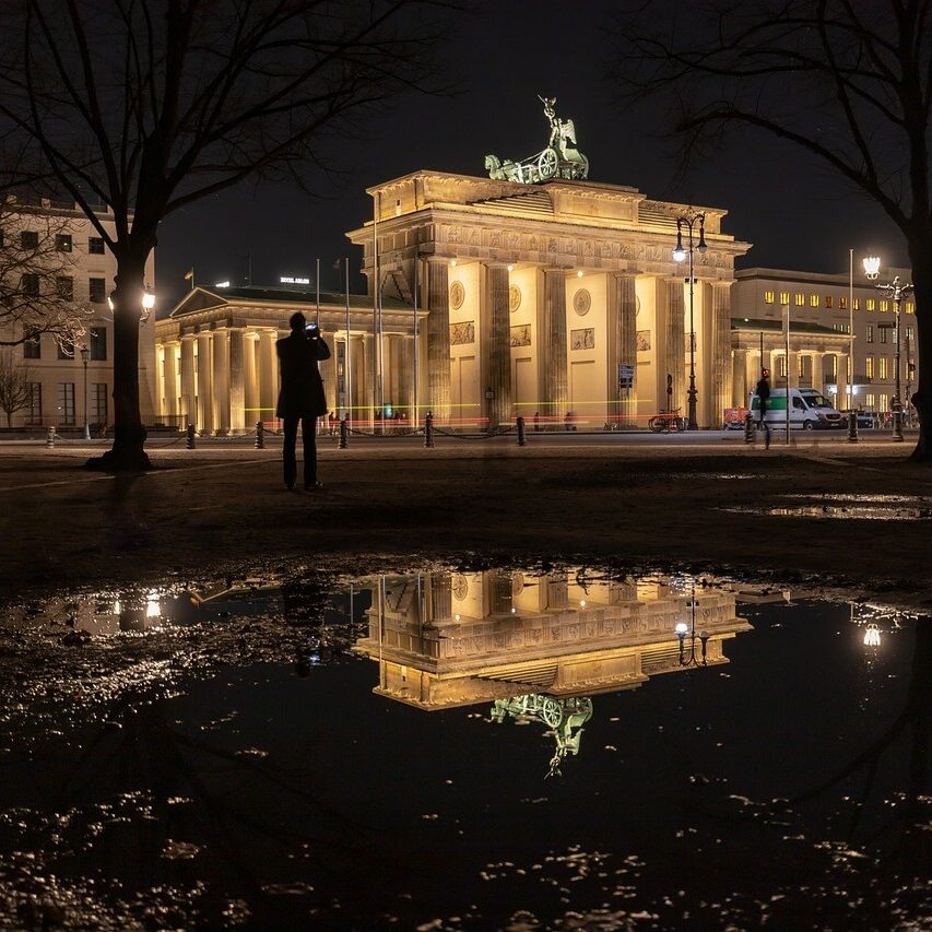 Das Nachtbild zeigt das Brandenburger Tor in Berlin, wunderschön beleuchtet und in einer großen Pfütze reflektiert. Silhouetten von Bäumen und einer einsamen Gestalt sind in der dunklen Umgebung subtil zu erkennen und verleihen der ruhigen Szene Tiefe.