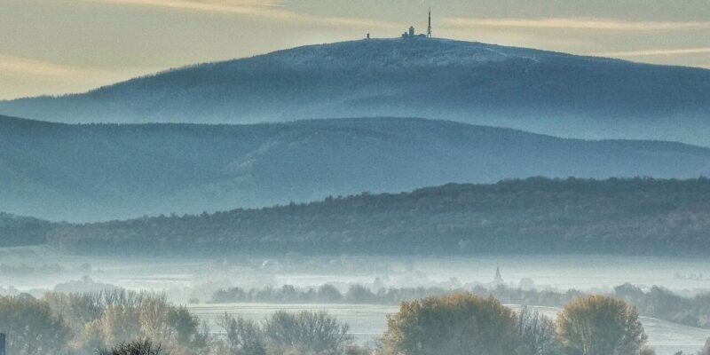 Eine ruhige Landschaft mit nebligen Hügeln, im Hintergrund der markante Brocken mit einem Gebäude und einer Antenne. Die Szene wird von Bäumen in Herbstfarben im Vordergrund eingerahmt, unter einem Himmel mit vereinzelten Wolken.