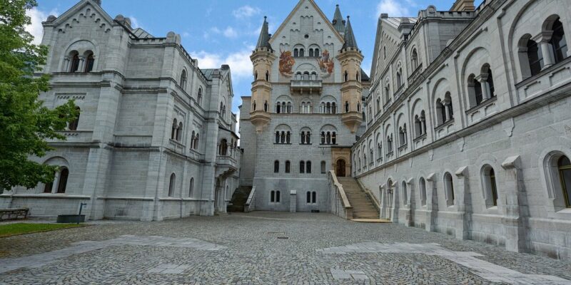 Ein Blick auf Schloss Neuschwanstein, ein prächtiges Steinschloss mit einem gepflasterten Innenhof. Das Schloss besticht durch hohe Türme, verzierte Fenster und komplizierte architektonische Details vor dem Hintergrund eines teilweise bewölkten Himmels.