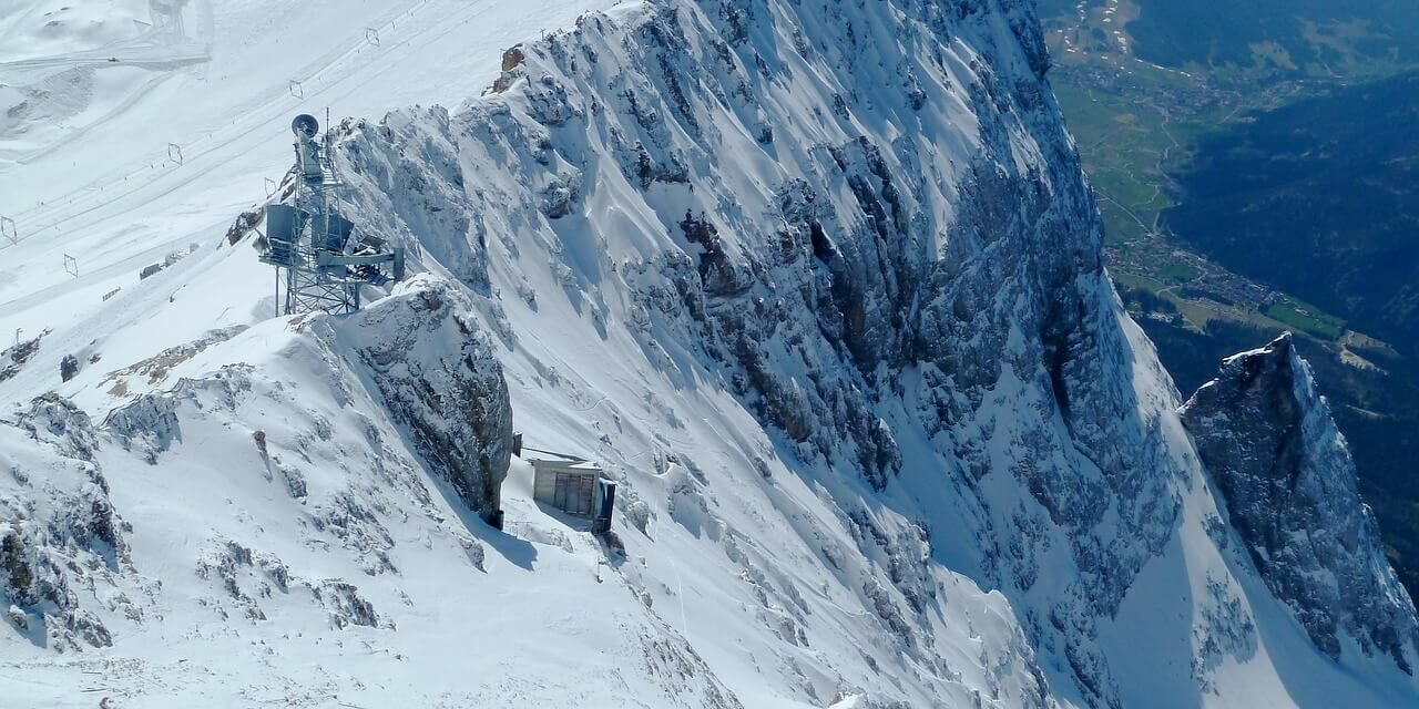 Schneebedeckter Berggipfel mit steilen Klippen und entfernten Tälern. Am Hang ist ein kleines Bauwerk zu erkennen, das von eisigem Gelände umgeben ist. Der Himmel ist klar und gibt den Blick frei auf die Berge und Wälder darunter, die in ihrer Erhabenheit an Deutschlands höchsten Berg erinnern.