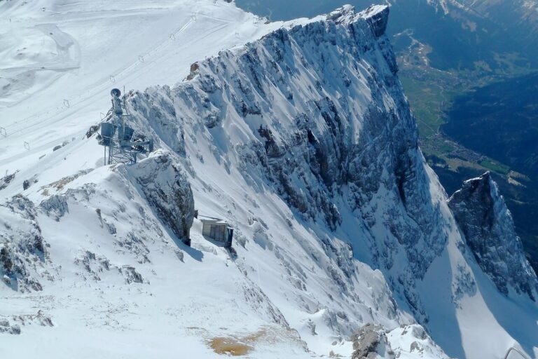 Schneebedeckter Berggipfel mit steilen Klippen und entfernten Tälern. Am Hang ist ein kleines Bauwerk zu erkennen, das von eisigem Gelände umgeben ist. Der Himmel ist klar und gibt den Blick frei auf die Berge und Wälder darunter, die in ihrer Erhabenheit an Deutschlands höchsten Berg erinnern.