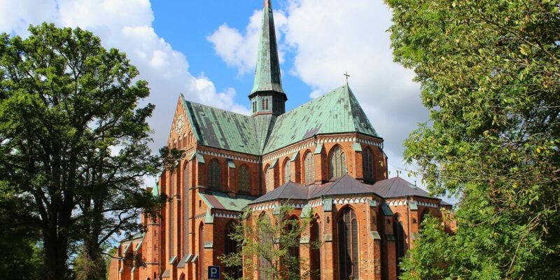 Doberaner Münster Das Doberaner Münster, eine große Kathedrale aus rotem Backstein mit grünem Kupferdach und hohem Turm, steht inmitten üppiger Bäume unter einem hellen, teilweise bewölkten Himmel. Seine detailreiche Architektur mit hohen Fenstern und gewölbten Türen erweckt einen majestätischen Eindruck. In der Nähe ist ein Parkschild zu sehen.