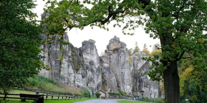 Ein malerischer Blick auf einen von Bäumen gesäumten Weg führt zu den hohen, schroffen Felsformationen der Externsteine im Teutoburger Wald. Der Weg ist von grünem Gras und einem Holzzaun gesäumt, wobei ein überhängender Baum einen natürlichen Rahmen in dieser bezaubernden Parklandschaft bildet.