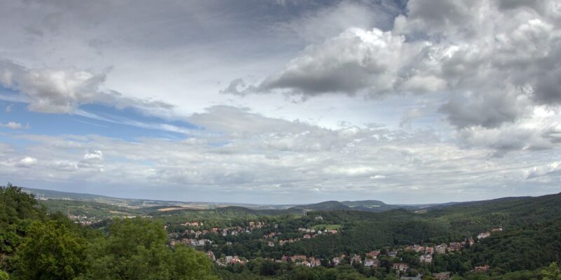 Unterhalb des majestätischen Großen Beerbergs eröffnet sich ein Panoramablick auf ein üppig grünes Tal mit verstreuten Häusern, die sich zwischen die Bäume schmiegen. Sanfte Hügel und ausgedehnte Wälder erstrecken sich in der Ferne unter einem teilweise bewölkten Himmel und schaffen eine ruhige und malerische Landschaft.