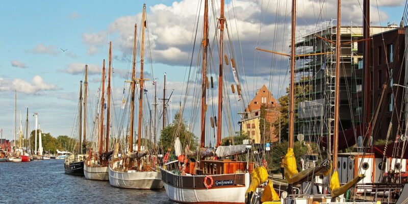 Eine Reihe von Segelbooten liegt am Flussufer der Hansestadt Greifswald. Der Himmel ist teilweise bewölkt und die gegenüberliegende Uferseite ist von Gebäuden gesäumt. Hohe Masten und Segel stechen ins Auge und schaffen eine friedliche Atmosphäre am Wasser, die diese charmante Stadt widerspiegelt.