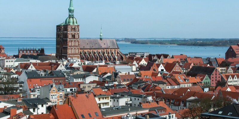 Ein malerischer Blick auf die historische Hansestadt Stralsund zeigt Gebäude mit roten Dächern und eine markante Kirche mit einem grünen Turm. Die Stadt ist wunderschön von einem großen Wasserkörper umgeben, der sich unter einem klaren blauen Himmel bis zum Horizont erstreckt.