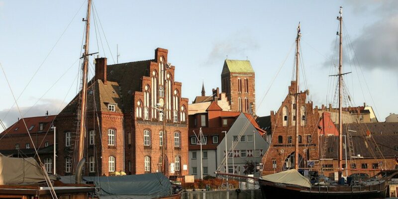 Hansestadt Wismar Ein malerischer Blick auf den Hafen der Hansestadt Wismar, mit traditionellen Backsteingebäuden im Hintergrund mit Stufengiebeln und einem hohen Kirchturm. Segelboote mit Masten liegen am Ufer vor Anker und spiegeln sich im ruhigen Wasser unter einem teilweise bewölkten Himmel.