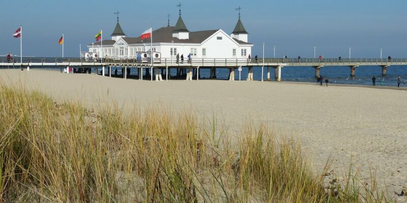 Insel Usedom Ein weißes Gebäude mit spitzen Dächern steht auf einem Pier über dem Meer auf der Insel Usedom. In der Nähe wehen mehrere Flaggen. Im Vordergrund ist ein Sandstrand mit hohen Gräsern unter einem klaren blauen Himmel zu sehen.