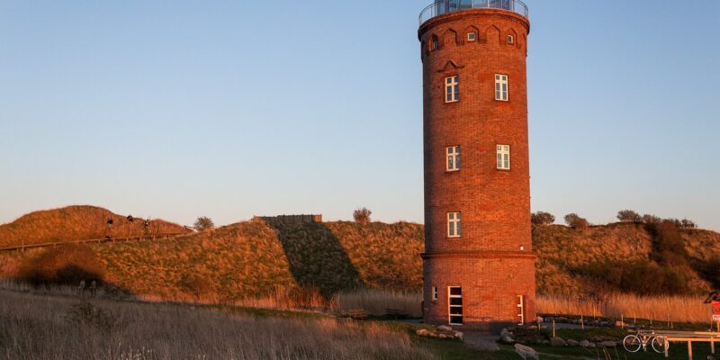 Kap Arkona Ein hoher, zylindrischer Backsteinturm, der an Kap Arkona erinnert, mit mehreren Fenstern und einer Glaskuppel oben drauf, steht auf einer Rasenfläche. Die Sonne wirft warmes, goldenes Licht auf den Turm und die umgebende Landschaft. In der Nähe seines Fußes sind ein Pfad und ein paar Fahrräder zu sehen.