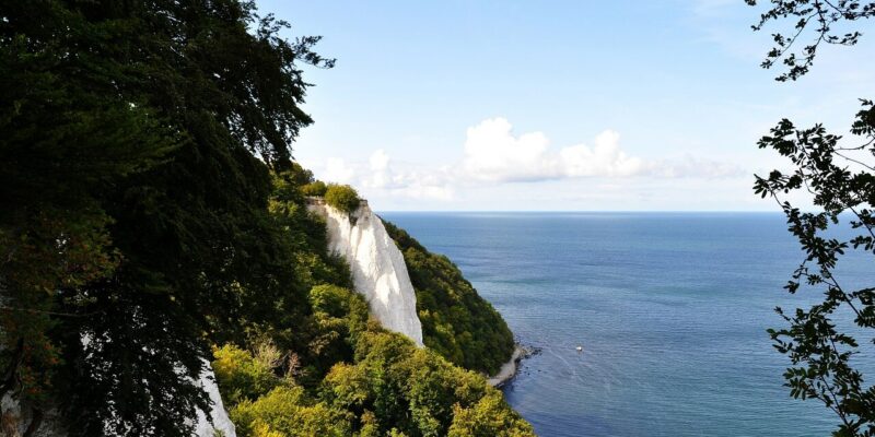Königsstuhl Malerischer Blick auf den majestätischen Königsstuhl, dessen weiße Kreidefelsen auf ein ruhiges, blaues Meer blicken. Üppige grüne Bäume rahmen die Klippen und das Meer ein, während ein klarer blauer Himmel mit ein paar Wolken diese ruhige Landschaft vervollständigt.