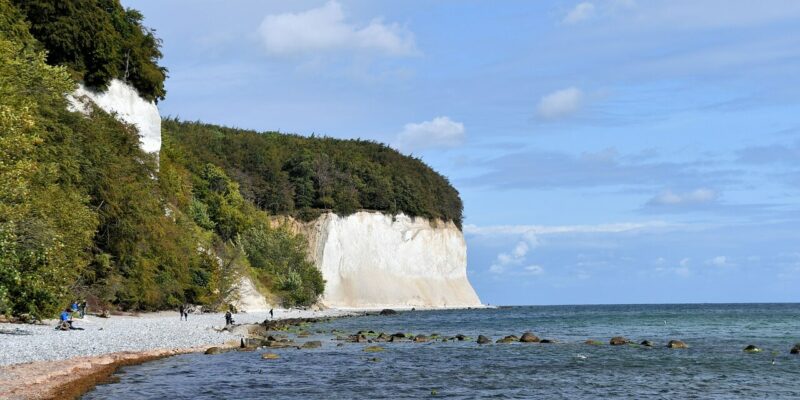 Kreidefelsen Küstenszene mit weißen Kreidefelsen, darunter die berühmten Kreidefelsen und der Königsstuhl, und einem Kieselstrand, der ans Meer grenzt. Mehrere Menschen gehen unter einem teilweise bewölkten Himmel am Ufer entlang, während üppige grüne Bäume die Klippen säumen.