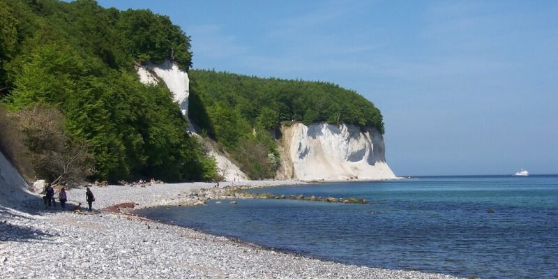 Ein felsiger Strand mit den majestätischen Kreidefelsen auf Rügen im Hintergrund, gesäumt von üppigen grünen Bäumen. Das ruhige blaue Meer erstreckt sich bis zum Horizont, wo ein kleines Boot unter einem klaren Himmel segelt. Mehrere Menschen schlendern am ruhigen Ufer entlang.