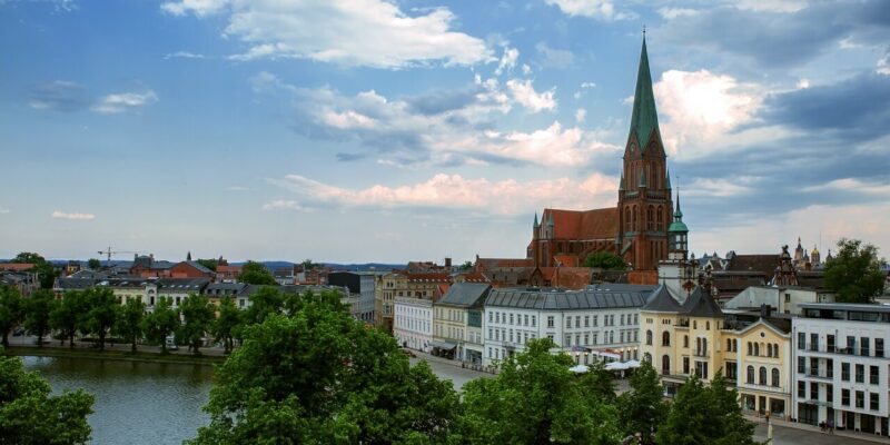 Eine malerische Aussicht auf die Landeshauptstadt Schwerin zeigt eine europäische Stadt mit einer großen Backsteinkirche mit einem hohen Turm, umgeben von historischen Gebäuden. Im Vordergrund ist der Fluss von üppigen grünen Bäumen unter einem bewölkten Himmel gesäumt.