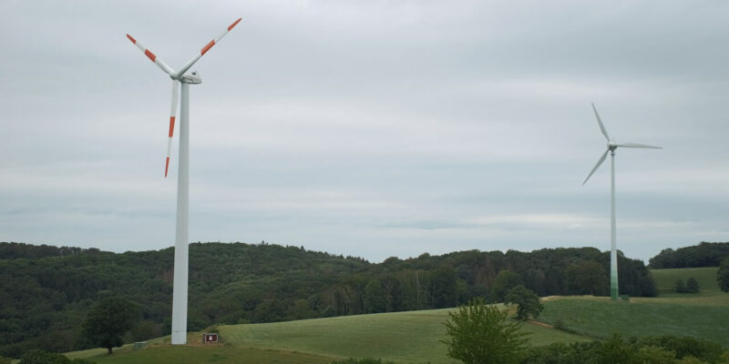 Zwei Windräder stehen auf den grünen Hügeln unter einem bedeckten Himmel. Die Landschaft besteht aus Grasfeldern und einem Waldgebiet im Hintergrund.