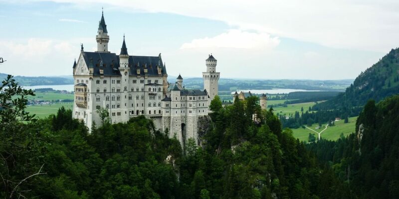Ein Märchenschloss mit hohen Türmen und Spitzen thront auf einem üppigen, grünen Hügel, umgeben von dichten Wäldern. Im Hintergrund erstreckt sich eine weite Landschaft mit Feldern und einem entfernten Gewässer unter einem teilweise bewölkten Himmel.