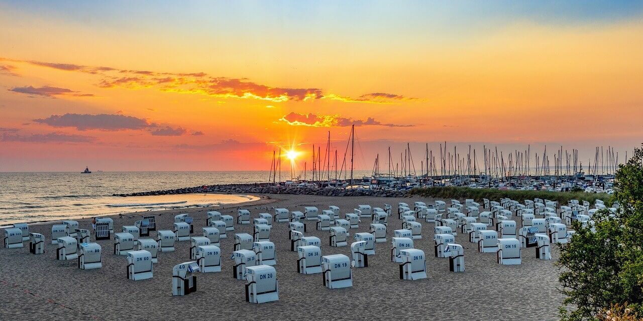 Mecklenburg-Vorpommern Sehenswürdigkeiten Ein malerischer Strand bei Sonnenuntergang mit Reihen weißer Stühle mit Blick aufs Meer fängt den Reiz der Sehenswürdigkeiten in Mecklenburg-Vorpommern ein. Der Himmel, eine leuchtende Mischung aus Orange und Lila, umrahmt Segelboote in der Nähe eines Wellenbrechers, während üppiges Grün den Sand säumt.