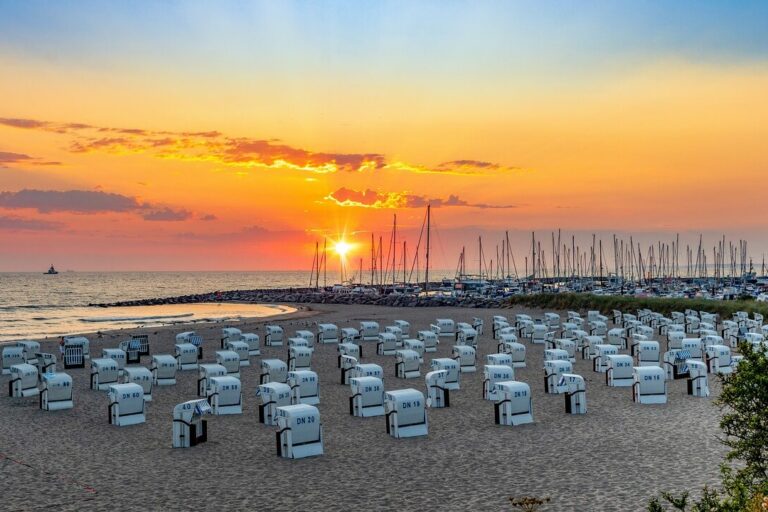 Mecklenburg-Vorpommern Sehenswürdigkeiten Ein malerischer Strand bei Sonnenuntergang mit Reihen weißer Stühle mit Blick aufs Meer fängt den Reiz der Sehenswürdigkeiten in Mecklenburg-Vorpommern ein. Der Himmel, eine leuchtende Mischung aus Orange und Lila, umrahmt Segelboote in der Nähe eines Wellenbrechers, während üppiges Grün den Sand säumt.