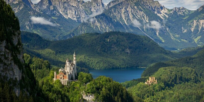 Ein malerischer Blick auf Schloss Neuschwanstein, eingebettet zwischen üppigen grünen Wäldern und einem See in der Nähe. Hohe Berge im Hintergrund unter einem teilweise bewölkten Himmel runden die malerische Landschaft ab.