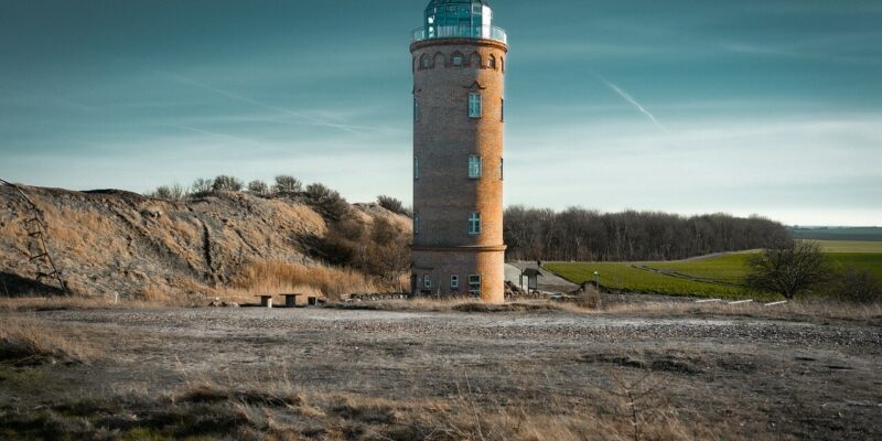 Peilturm Kap Arkona Ein hoher, zylindrischer Leuchtturm namens Peilturm Kap Arkona steht in einer kargen Landschaft mit spärlichem Gras. Der Himmel ist klar mit ein paar dünnen Wolken, und in der Ferne ist am Horizont eine Reihe von Bäumen zu erkennen.