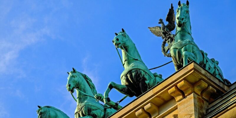 Eine Nahaufnahme der Quadriga auf dem Brandenburger Tor in Berlin, die einen von vier Pferden gezogenen Wagen vor einem klaren blauen Himmel zeigt. Die detailliert gearbeiteten Bronzefiguren wirken majestätisch und markant und dominieren die Szene mit ihrer Erhabenheit.