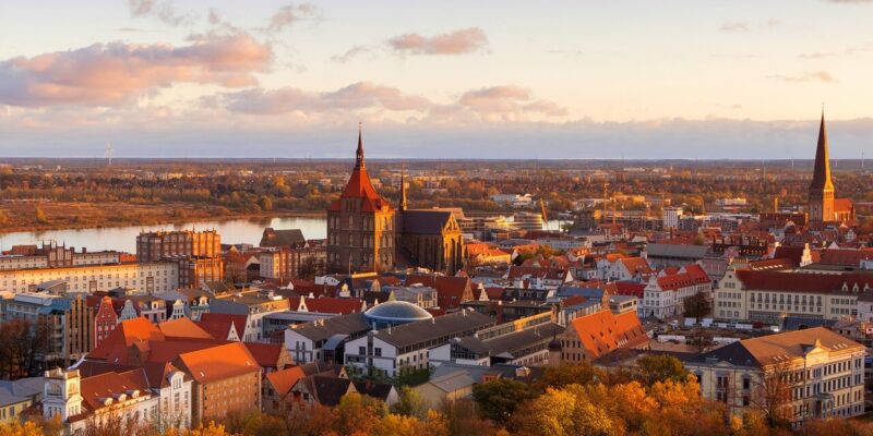Luftaufnahme der historischen Stadt Rostock mit rot gedeckten Gebäuden, einer markanten Kirche mit hohem Turm und einem Fluss im Hintergrund. Die Bäume in der Umgebung zeigen sich in leuchtenden Herbstfarben unter einem teilweise bewölkten Himmel.
