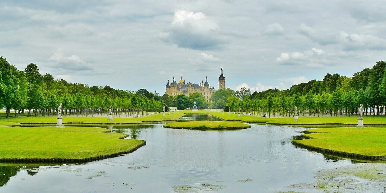 Schloss Schwerin, ein prachtvolles Schloss mit spitzen Türmen, liegt in der Ferne, umgeben von üppigem Grün. Im Vordergrund spiegelt ein ruhiger Teich den bewölkten Himmel wider, gesäumt von gepflegten Rasenflächen und Baumreihen. Statuen schmücken beide Seiten des Wassers.