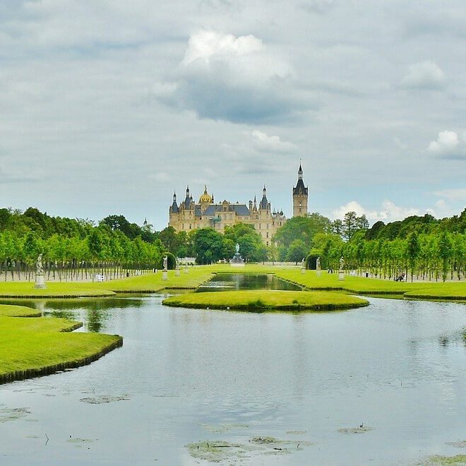 Schloss Schwerin, ein prachtvolles Schloss mit spitzen Türmen, liegt in der Ferne, umgeben von üppigem Grün. Im Vordergrund spiegelt ein ruhiger Teich den bewölkten Himmel wider, gesäumt von gepflegten Rasenflächen und Baumreihen. Statuen schmücken beide Seiten des Wassers.