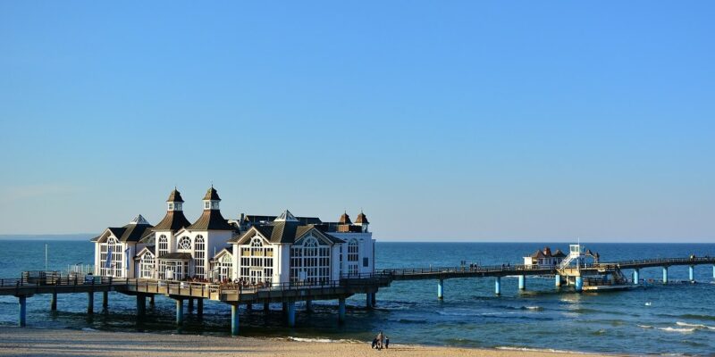 Seebrücke Sellin Eine malerische Anlegestelle, die an die Seebrücke Sellin erinnert, erstreckt sich in das ruhige blaue Meer und verfügt über ein charmantes Gebäude mit mehreren Spitzdächern. Am Sandstrand im Vordergrund sind einige Menschen zu sehen, die einen sonnigen Tag unter klarem Himmel genießen.