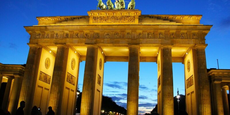 Das Bild zeigt das beleuchtete Brandenburger Tor in der Abenddämmerung. Der Himmel ist tiefblau und im Vordergrund sind Silhouetten von Menschen zu sehen. Die Details des Tors und die Statuen heben sich vom Abendhimmel ab.