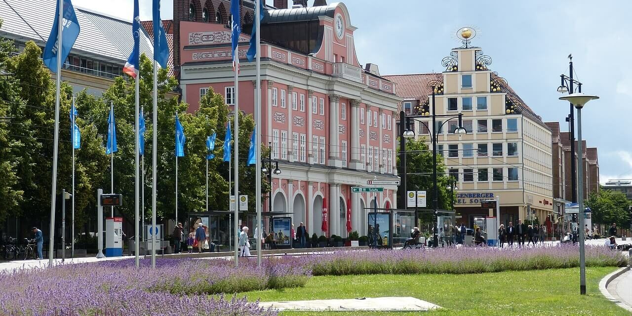 Eine lebhafte Straßenszene, typisch für Städte in Mecklenburg-Vorpommern, entfaltet sich mit einem Rasen und violetten Blumen im Vordergrund. Blaue Flaggen säumen die Straße, während bunte Gebäude, darunter ein rosafarbenes mit Bogenfenstern, Charme verleihen. Menschen schlendern unter dem bewölkten Himmel.