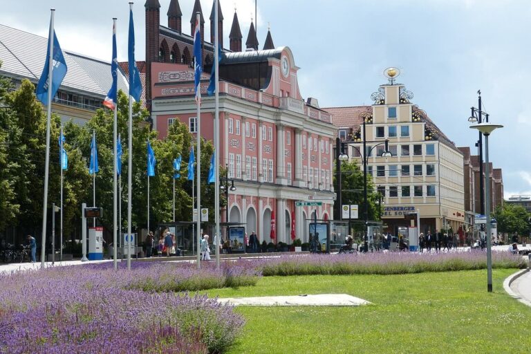 Städte in Mecklenburg-Vorpommern Eine lebhafte Straßenszene, typisch für Städte in Mecklenburg-Vorpommern, entfaltet sich mit einem Rasen und violetten Blumen im Vordergrund. Blaue Flaggen säumen die Straße, während bunte Gebäude, darunter ein rosafarbenes mit Bogenfenstern, Charme verleihen. Menschen schlendern unter dem bewölkten Himmel.