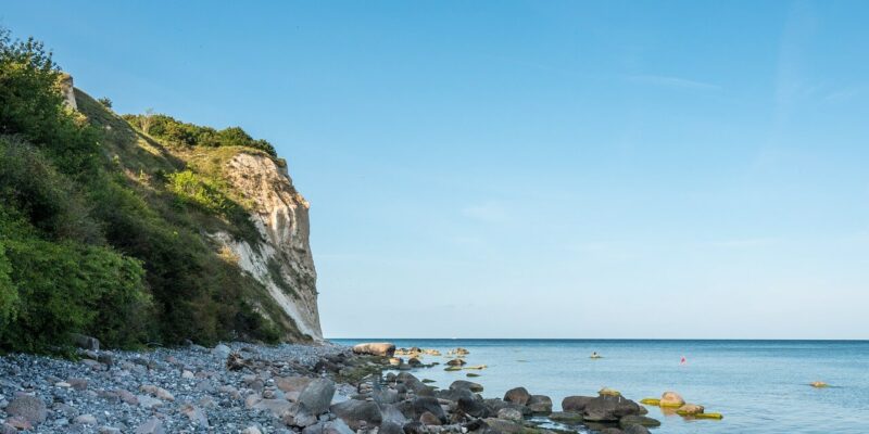 Steilküste Kap Arkona auf der Insel Rügen Eine Küstenszene an der Steilküste Kap Arkona auf der Insel Rügen zeigt einen felsigen Strand mit verstreuten Steinen, der zu einer steilen, grasbewachsenen Klippe auf der linken Seite hinaufführt. Das ruhige blaue Meer erstreckt sich unter einem klaren blauen Himmel bis zum Horizont.