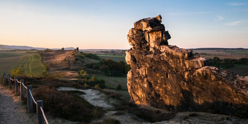 Die Teufelsmauer im Harz ist eine beeindruckende große Felsformation in einer hügeligen Landschaft, getaucht in das warme Licht des Sonnenuntergangs. Ein von einem Holzzaun gesäumter Feldweg schlängelt sich entlang der sanften Hänge, während der klare Himmel einen sanften Farbverlauf bietet, der die bezaubernde Aussicht noch verstärkt.
