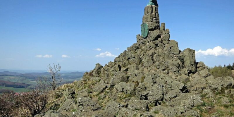 Felsmonument auf der Wasserkuppe mit einer Vogelskulptur auf der Spitze, vor einem klaren blauen Himmel. Im Vordergrund sind unebene Felsen zu sehen, die den Blick auf Felder und Wälder bis zum Horizont freigeben. Spärliche Vegetation klammert sich an die schroffe Struktur.