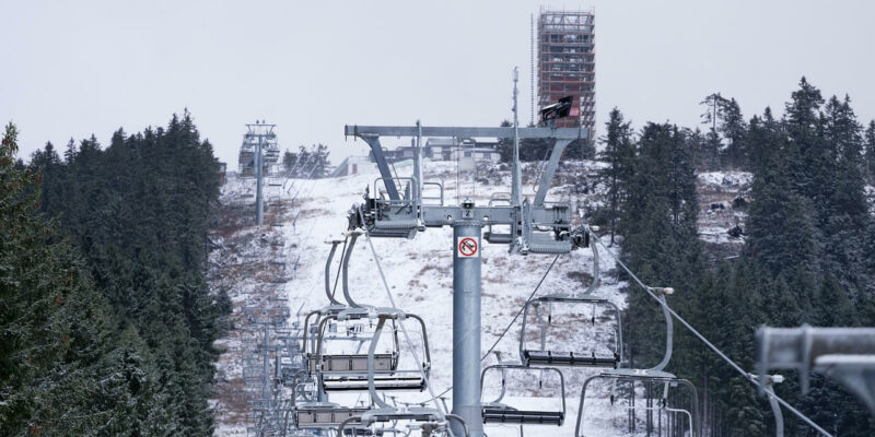 Ein Skilift mit leeren Sesseln fährt den schneebedeckten Wurmberg hinauf, umgeben von dichten, dunklen immergrünen Bäumen. Im Hintergrund wird vor einem wolkigen Himmel ein hohes Bauwerk errichtet.