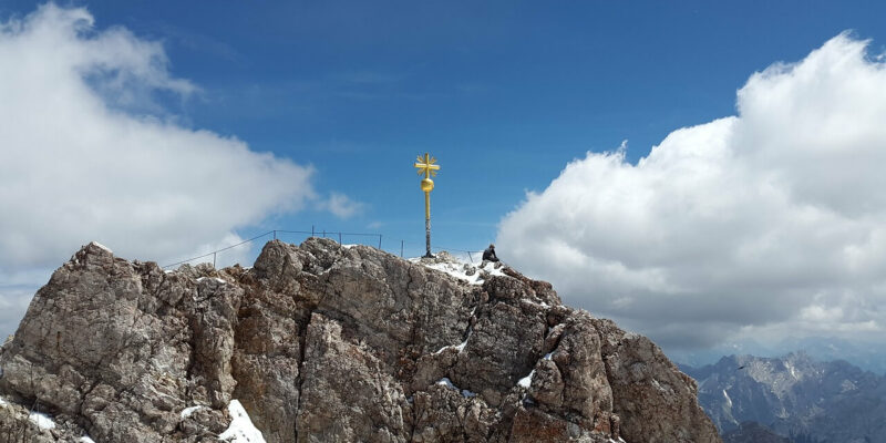 Ein felsiger Berggipfel, der an die Zugspitze erinnert, zeigt ein leuchtend gelbes Kreuz auf dem Gipfel unter einem klaren blauen Himmel mit vereinzelten Wolken. Auf der zerklüfteten Oberfläche sind Schneeflecken sichtbar und die Berge in der Ferne bilden eine beeindruckende Kulisse.