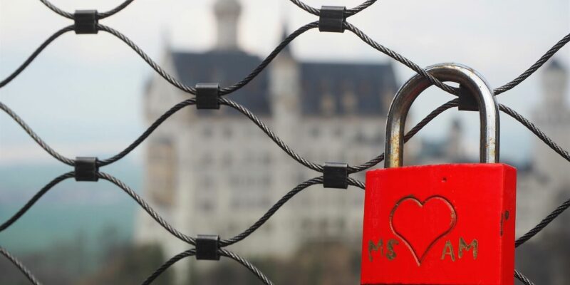 An einem Maschendrahtzaun ist ein rotes Vorhängeschloss mit einem Herz und den Initialen MS und AM befestigt. Im unscharfen Hintergrund ist vor einem wolkigen Himmel ein Schloss mit hohen Türmen zu erkennen.