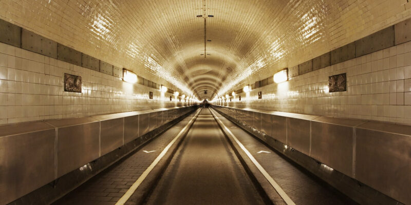 Der Alte Elbtunnel in Hamburg besteht aus einem langen, symmetrischen Durchgang mit weiß gefliesten Wänden und einer Decke, die von Lichtreihen beleuchtet wird. Die leere Straße im Inneren zeigt sichtbare Fahrspuren und schwache Reflexionen, während der Tunnel in die Ferne verschwindet und einen faszinierenden Fluchtpunkteffekt erzeugt.