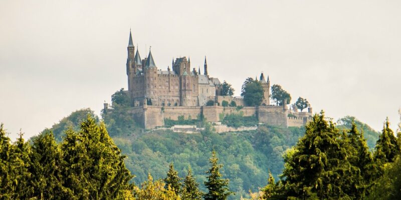 Burg Hohenzollern, eine mittelalterliche Burg auf einem bewaldeten Hügel, ist von üppigem Grün umgeben. Ihre Türme und Türmchen ragen vor einem wolkigen Himmel empor und bilden eine ruhige und majestätische Landschaft.