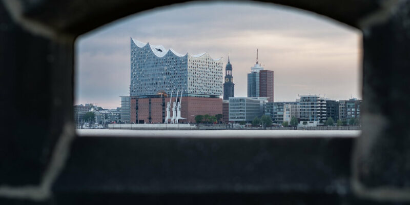 Werfen Sie einen Blick auf die Elbphilharmonie in Hamburg, perfekt eingerahmt durch eine Steinöffnung. Die moderne Glasfassade bildet einen schönen Kontrast zu den historischen Gebäuden unter einem stimmungsvollen, wolkigen Himmel.
