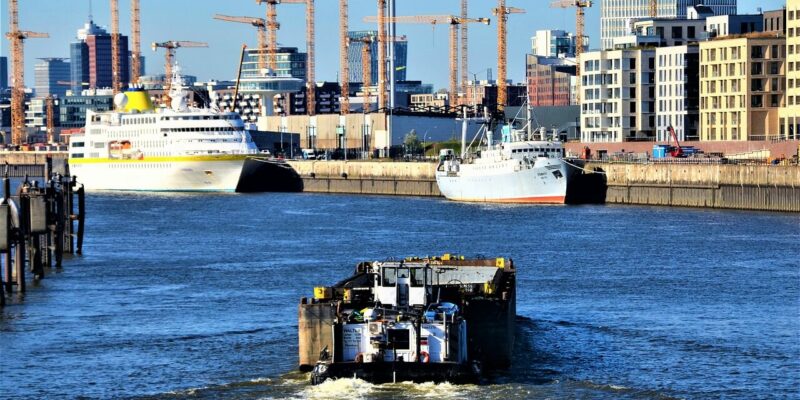 Ein Frachtschiff fährt auf einem Fluss auf zwei große angedockte Schiffe zu. Im Hintergrund ist die Skyline der HafenCity in Hamburg mit zahlreichen Baukränen und modernen Gebäuden gesäumt, die sich unter einem klaren blauen Himmel am Wasser entlang erstrecken.
