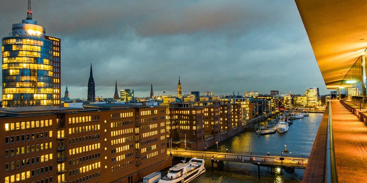 Eine Stadtlandschaft in der Abenddämmerung mit beleuchteten Bürogebäuden entlang eines Kanals zeigt einige der Sehenswürdigkeiten Hamburgs. Boote liegen entlang des Wasserwegs vor Anker und eine Brücke überquert den Kanal. Der bewölkte Himmel fängt einen warmen Schein der Lichter der Stadt ein, die sich im Wasser spiegeln.