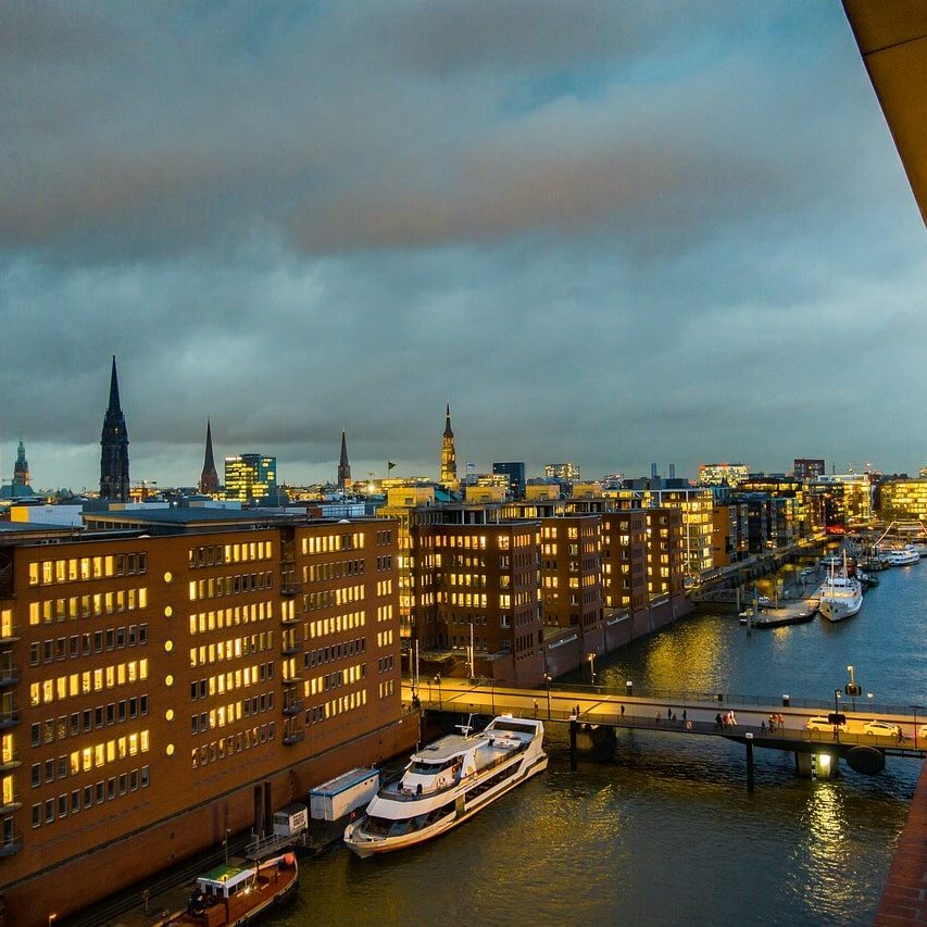 Eine Stadtlandschaft in der Abenddämmerung mit beleuchteten Bürogebäuden entlang eines Kanals zeigt einige der Sehenswürdigkeiten Hamburgs. Boote liegen entlang des Wasserwegs vor Anker und eine Brücke überquert den Kanal. Der bewölkte Himmel fängt einen warmen Schein der Lichter der Stadt ein, die sich im Wasser spiegeln.
